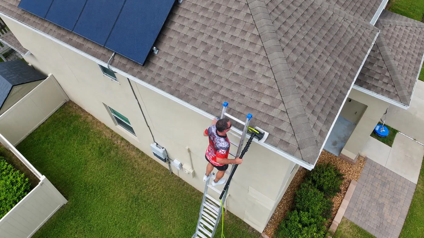 Iron Shield technician working from an extension ladder with a long-reach tool, clearing buildup from the gutter and roofline