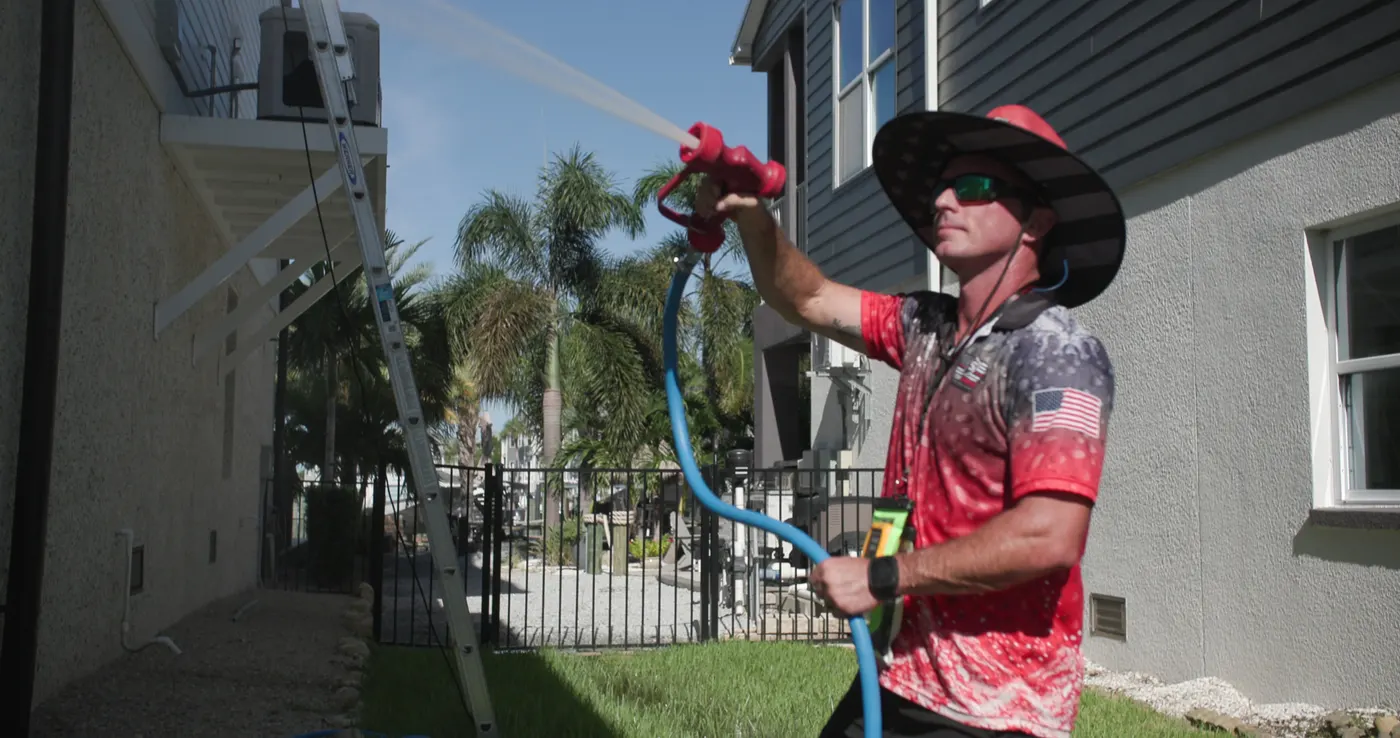Iron Shield technician in a wide-brimmed hat aiming a soft wash spray gun up at the exterior wall of a two-storey home