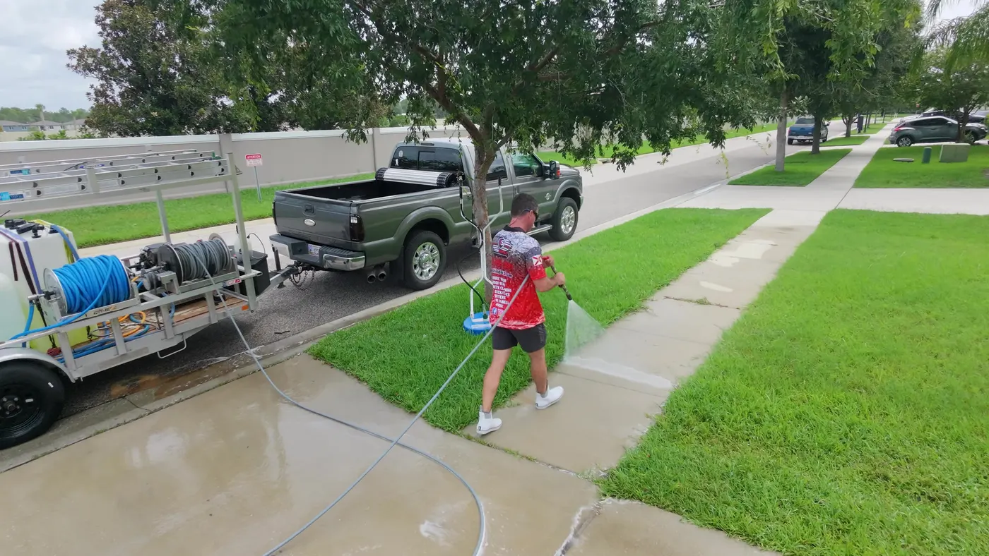 Aerial view of an Iron Shield technician pressure washing a concrete sidewalk, the cleaned section wet and brighter, the truck and equipment trailer at the curb