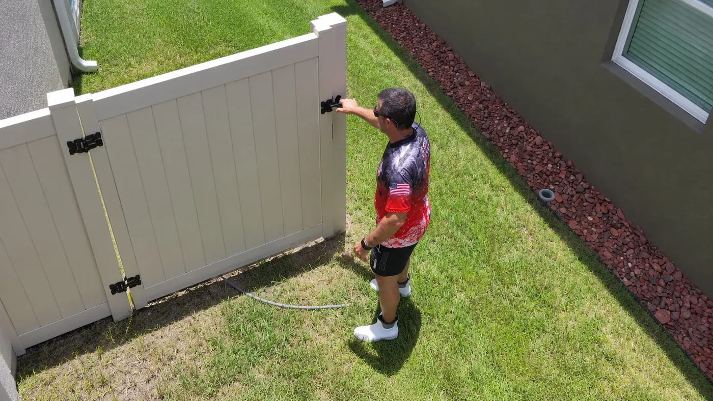 Iron Shield technician rinsing a white vinyl fence panel above a red mulch bed beside a Hudson home