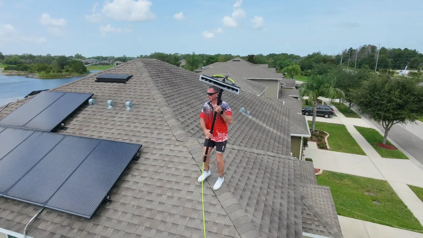 Iron Shield technician using a long-handled brush on a shingle roof next to a solar panel array