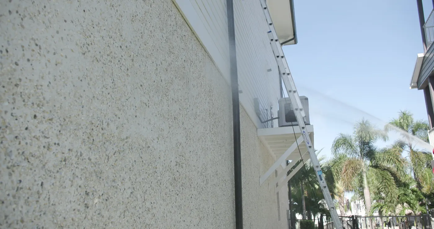 Low-pressure spray arcing up a tall textured masonry wall, a ladder set against the side of the home