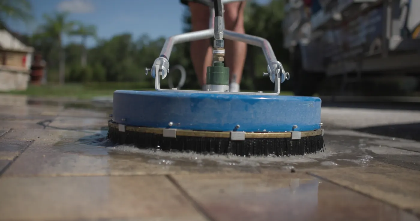 Close-up of a rotary surface cleaner moving across wet pavers, its brush skirt holding the spray to the surface