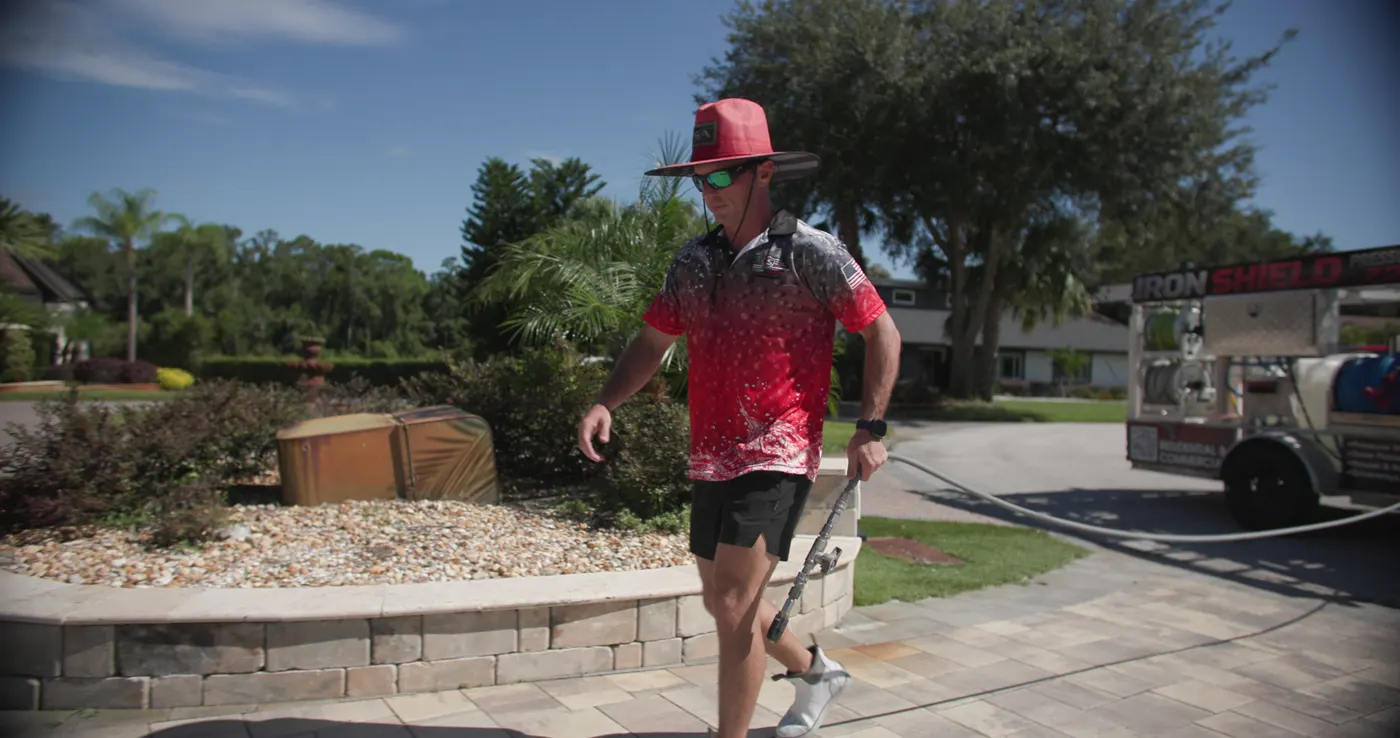 Iron Shield technician carrying a spray wand across a paver walkway beside a stone-block planter wall, the service trailer parked behind
